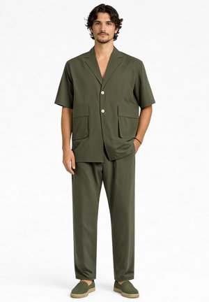 Mężczyzna stoi, ubrany w oliwkowo-zieloną, krótki rękaw, zapinaną na guziki kurtkę z dużymi kieszeniami, pasujące spodnie i buty wsuwane na tle jednolitego tła.