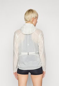 Lightweight, translucent jacket in white with a fitted hood, a zipper pocket, and reflective details along the back. Smooth texture.