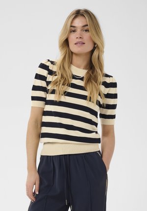 Femme aux longs cheveux blonds porte un haut en maille rayé crème et noir à manches courtes et un pantalon bleu marine avec cordon, les mains dans les poches.