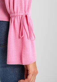 Maglione rosa lavorato a maglia con maniche a campana, con un laccio stretto al polso. Il materiale sembra morbido e dalla texture liscia. Indossato sopra un denim.