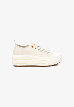 Zapatilla beige de lona de corte bajo con suela blanca gruesa, cordones blancos, ojal naranja cerca de la parte superior y lengüeta en el talón.