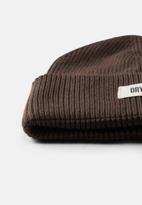 Gorro de malha canelada castanho com a bainha dobrada e um rótulo de tecido branco com o texto "DRY". Textura macia e forma estruturada.