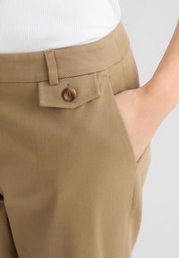 Pantaloni khaki con una texture liscia, dotati di una tasca frontale con un dettaglio di bottone marrone e una vestibilità slim in vita.
