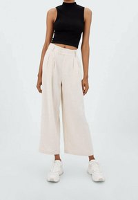 Linnen wijde-leg broek in lichtbeige, met een hoge taille voorzien van plooien en twee knopen aan de voorkant. Gecombineerd met een zwart geribd crop top en witte sneakers.