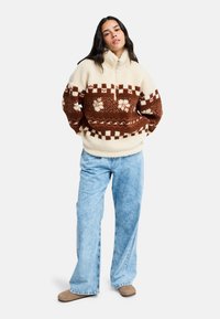 Pullover in pile color crema con motivi geometrici e floreali marroni. Presenta un colletto alto e una vestibilità comoda, abbinato a jeans a gamba larga color azzurro chiaro.