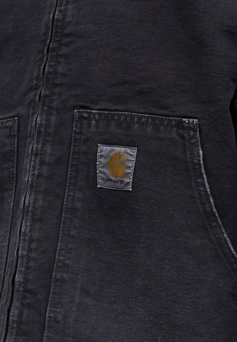 Nahaufnahme der Tasche einer dunkelgrauen Stoffjacke mit einem aufgenähten Carhartt-Logo-Patch in Gelb und Rot.