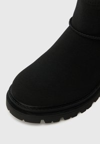 Bota de tornozelo em camurça preta com bico arredondado, sola de borracha plana e detalhes cosidos ao longo da costura superior.