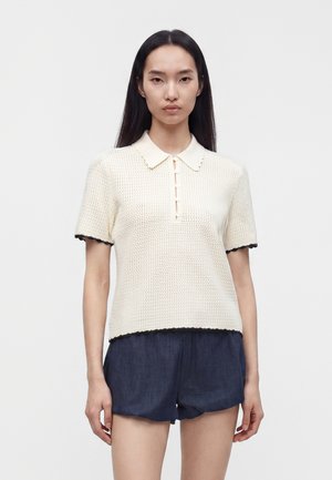 Jeune femme aux cheveux noirs et raides porte un polo en maille crème à manches courtes avec bordure noire et un short bleu foncé, debout sur un fond uni.