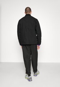 Uomo visto di spalle che indossa una giacca e pantaloni neri, con i polsini di un maglione viola visibili, scarpe Salomon grigie e blu, in piedi su uno sfondo semplice.
