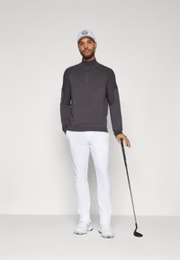 Dunkelgrauer Quarter-Zip-Pullover mit strukturierten Ärmeln, kombiniert mit weißen Hosen und weißen Golfschuhen. Hält einen Golfschläger.