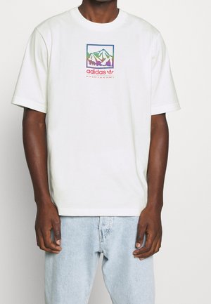 Homme portant un t-shirt Adidas blanc avec un graphique de montagne multicolore et des coordonnées, associé à un jean bleu clair, sur un fond uni.