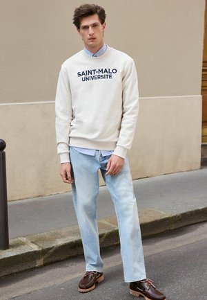 Felpa crema con collo rotondo e scritta blu "SAINT-MALO UNIVERSITÉ", indossata sopra una camicia a righe blu. Jeans blu chiaro e mocassini marroni.