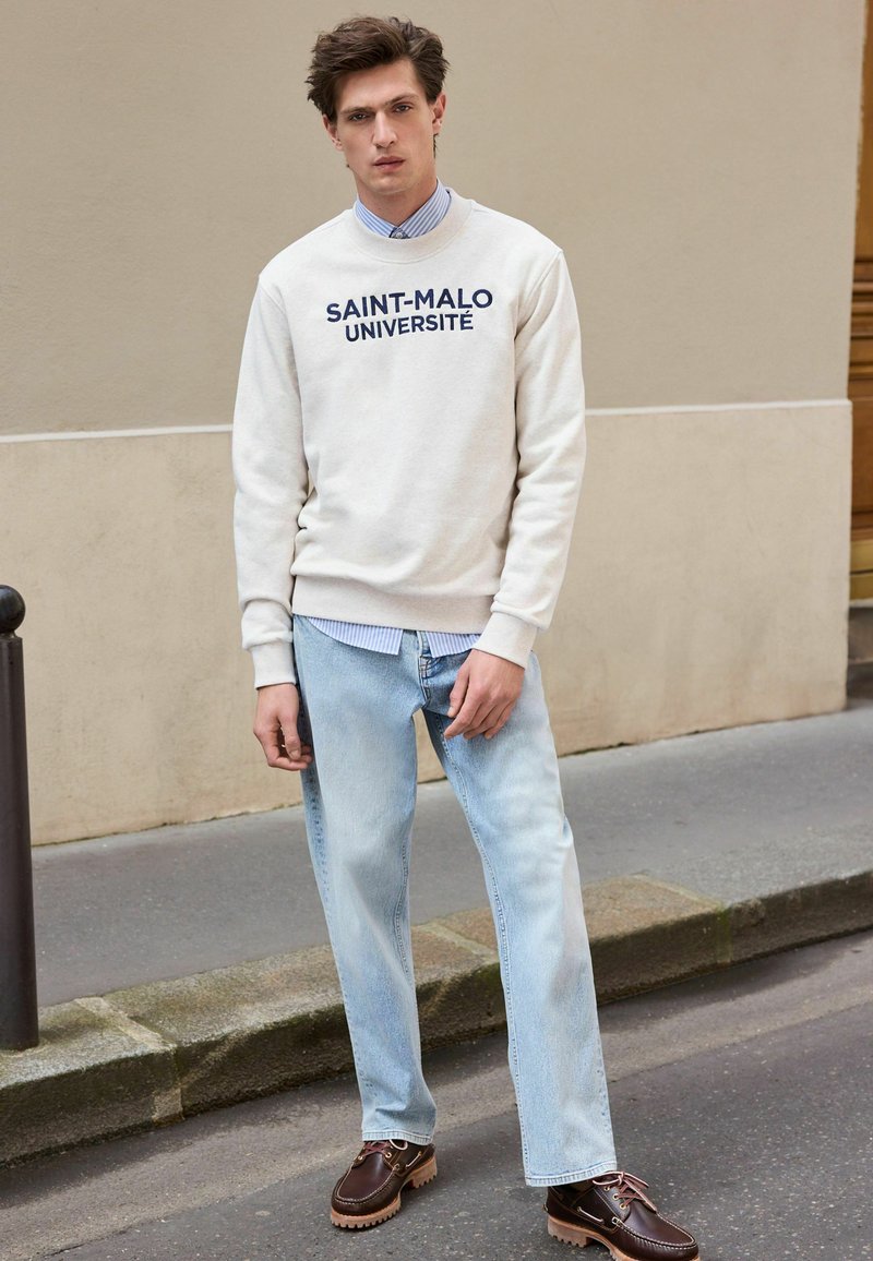 Lys beige sweatshirt med rund hals og marineblå tekst "SAINT-MALO UNIVERSITÉ," lagd over en blåstribet skjorte. Lyst blå jeans og brune loafers.