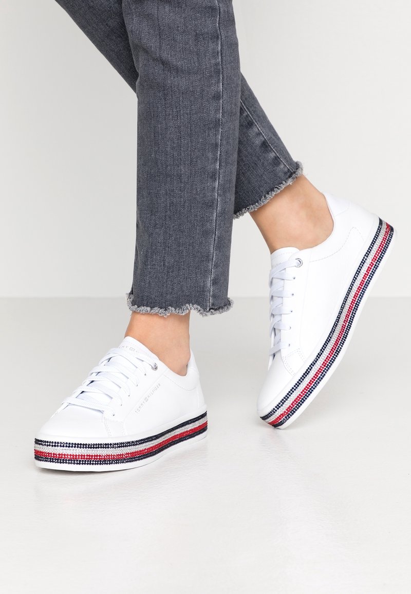 Białe sneakersy z płaską podeszwą, zdobione kolorowymi paskami w czerwonym, białym i niebieskim kolorze wzdłuż dolnej krawędzi. Cholewka z teksturowanej skóry.