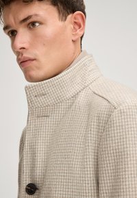 Beige jas met houndstoothpatroon, hoge kraag, knoopsluiting enDetails van textuur in de stof. Gladde voering zichtbaar bij de kraag.