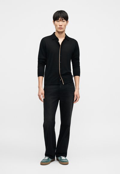 Μαύρο ζακέτα με κουμπιά και μακριά μανίκια, με αντίθετη μπεζ ρίγα, σε συνδυασμό με σκούρο τζιν και πετρόλ αθλητικά παπούτσια. Απλός, καθημερινός σχεδιασμός.