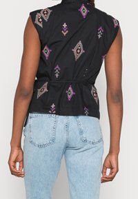 Persona che indossa una canotta nera senza maniche con ricami geometrici multicolori e jeans a vita alta blu chiaro, vista di spalle.