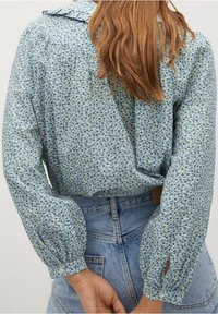 Blaue Bluse mit floralem Muster, gerafften Schulterdetails, Puffärmeln und Kragen. Der Stoff ist leicht, mit einer sanften Textur und einem filigranen Muster.