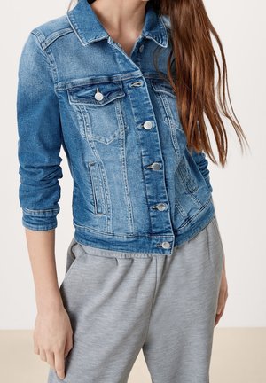 Vrouw draagt een nauwsluitend blauwe spijkerjasje met zilveren knopen en grijze casual broek, met lang bruin haar over de schouder.