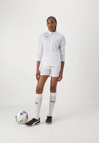 Chaqueta deportiva blanca con cremallera, pantalones cortos a juego, botas de fútbol negras, calcetas blancas hasta la rodilla con logotipo y un balón de fútbol multicolor en el suelo.