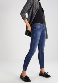 Jeans skinny de tiro medio en denim azul oscuro con una textura suave y un ligero desgaste. Combinados con una blusa negra y una americana gris texturada.