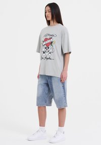 Lichtgrijze oversized t-shirt met een schedel- en hartgrafiek, gecombineerd met knielange vervaagde denim shorts en witte sneakers, staande tegen een witte achtergrond.
