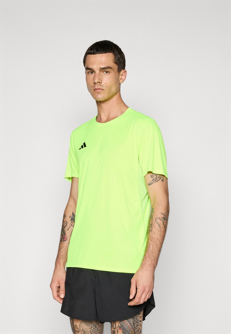 Man in een neon groene sport T-shirt met korte mouwen en zwarte shorts, staand tegen een effen grijze achtergrond, met zichtbare tatoeages op armen en benen.