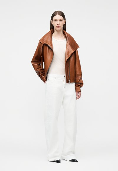 Γυναίκα με ίσια καστανά μαλλιά φοράει καφέ δερμάτινο μπουφάν με υπερμεγέθη γιακά, λευκό t-shirt, φαρδιά λευκή παντελόνια και μαύρα μυτερά παπούτσια.