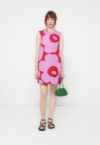 Rochie scurtă de un roșu vibrant, cu modele mari de flori roz, realizată dintr-un material neted. Asortată cu o geantă mică verde și sandale negre.