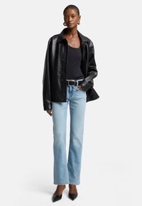 Giacca camicia oversize in pelle nera sopra un top aderente nero, abbinata a jeans svasati azzurri chiari e tacchi a punta neri.