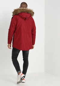 Abrigo parka rojo con capucha forrada de piel, que cuenta con cintura con cordón ajustable, mangas largas y una parte trasera ligeramente alargada. Combinado con vaqueros oscuros y zapatillas.