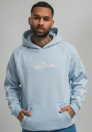 Man with neck and hand tattoos wearing light blue hoodie with "dada" logo and zippered front pockets, standing against plain background.