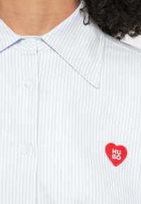 Svetlomodrá prúžkovaná košeľa na gombíky, s červenou nášivkou v tvare srdca s nápisom "HUGO". Tkanina vyzerá hladko s klasickým golierom.
