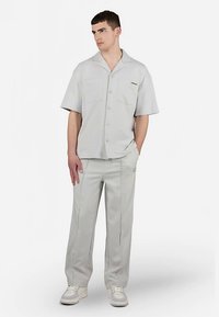 Νεαρός άνδρας με ανοιχτό γκρι κοντομάνικο πουκάμισο, ταιριαστά παντελόνια με ραφές, και λευκά αθλητικά παπούτσια, στέκεται με το ένα χέρι στην τσέπη.