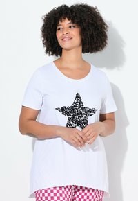 Hvid t-shirt med korte ærmer og et stort sort pailletten-stjerne design foran samt en rund halsudskæring, parret med lyserøde ternede bukser.