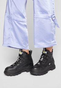 Botines negros hasta el tobillo con cuero texturizado, suelas gruesas de goma y cordones blancos. Combinados con pantalones cortos de color púrpura claro.