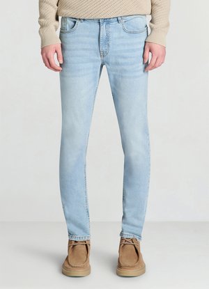 Jean skinny bleu clair avec une finition délavée, conception à cinq poches et couture contrastante. Le modèle est porté avec des chaussures en daim marron.