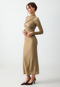 Robe dorée à manches longues avec des détails froncés et un col haut. Tissu lisse et légèrement brillant tombant jusqu'à la cheville. Associée à des talons effilés foncés.