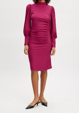 Eng anliegendes magentafarbenes Kleid mit langen Puffärmeln, gerippter Textur, hohem Kragen und seitlichen Raffdetails. Kombiniert mit spitzen schwarzen High Heels.