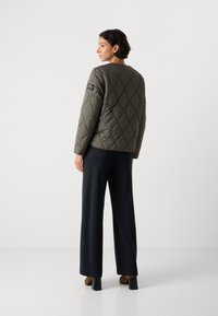 Vrouw met kort zwart haar, gekleed in een olijfgroene gewatteerde jas, zwarte wijde broek en bruine hakken, staand tegen een effen witte achtergrond.