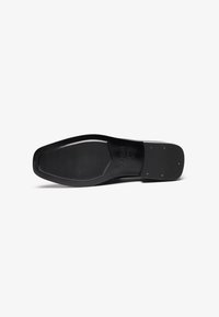 Suela de zapato de cuero negro con forma rectangular, que presenta una superficie texturada y pequeños tachuelas doradas cerca del talón para mejorar el agarre.