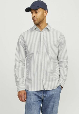 Camisa de botón a rayas en tejido ligero con rayas verticales en azul claro y blanco, con un bolsillo en el pecho. Combinada con jeans de mezclilla y una gorra azul marino.