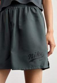 Nike šortky v tmavě šedé barvě, vyrobené z hladkého, lehkého materiálu. Mají elastický pas a výrazné černé logo na dolní části nohavice.