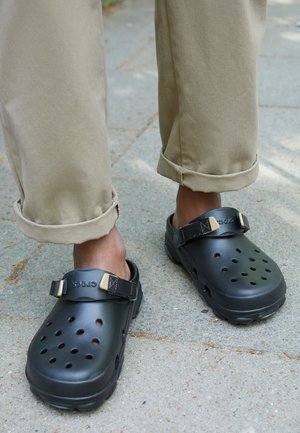 Des chaussures noires de style sabot avec des trous de ventilation, une sangle ajustable et une finition texturée, portées avec un pantalon kaki retroussé sur une surface en béton.