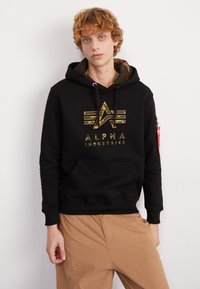 Felpa nera con cappuccio e foderatura interna in camouflage, dotata di un logo camo dorato e tasca frontale. Maniche lunghe con polsini a coste.