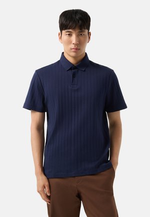 Mand iført en marineblå kortærmet ribbet poloshirt og brune bukser står mod en ensfarvet lys baggrund.