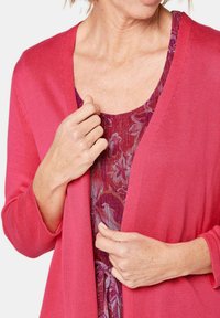 Roze vest met een getextureerd breiontwerp, gedragen over een bloemenprint jurk in verschillende tinten paars en rood. Korte mouwen en open voorkant.