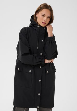 Woman wearing a long black hooded coat with snap buttons and large front pockets, posing with one hand near her face and crossed arms.
