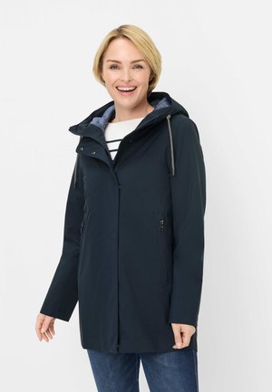 Lächelnde Frau trägt eine dunkelblaue Kapuzenjacke mit Reißverschlusstaschen über einem weißen gestreiften Hemd und blauer Jeans vor einem schlichten Hintergrund.