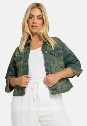 Lyshåret kvinde iført en falmet grøn denimjakke over en hvid top og hvide bukser med snor, hænder holder jakkeenderne.